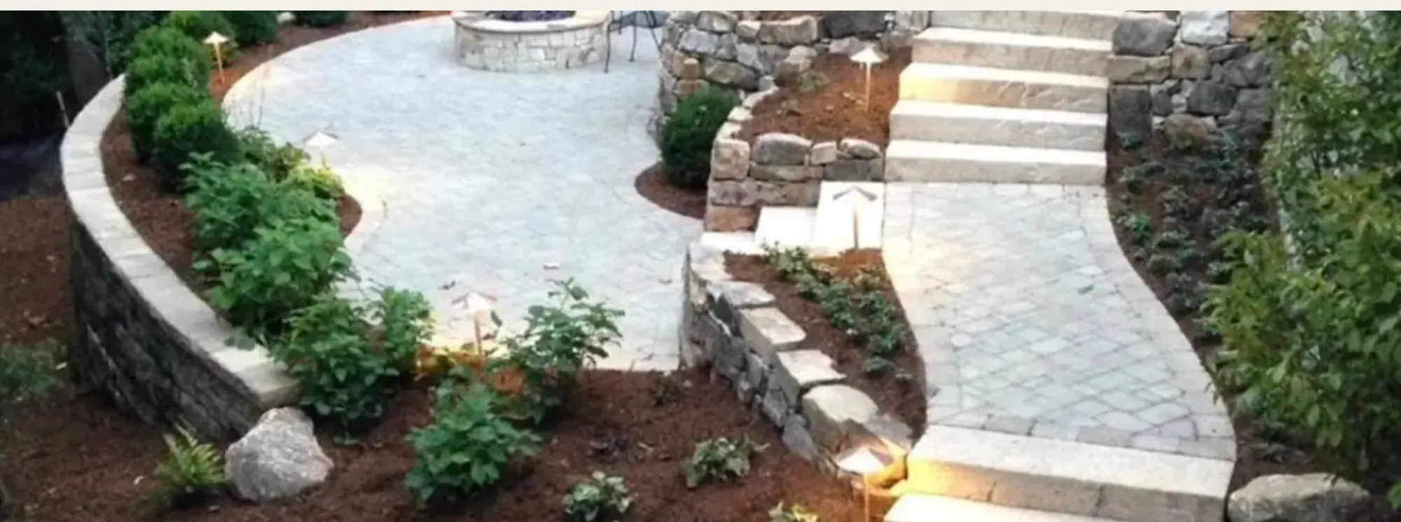 A completed Visionary Construction project on the live site — a steep rear yard rebuilt as a curved paver terrace with a stone fire pit, granite stair treads climbing the grade, and boulder-set retaining walls lit at dusk.